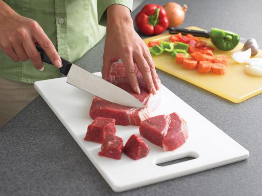 Someone cutting raw beef on a cutting board, with a separate cutting board with chopped up carrots, peppers, and onions to the side.